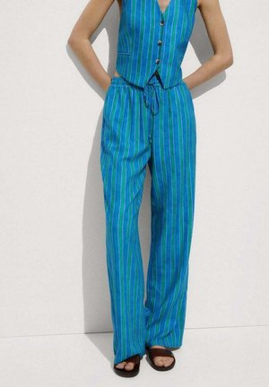 Persona que lleva un chaleco sin mangas con rayas verticales azules y verdes y pantalones de pierna ancha a juego con cordón, combinados con sandalias marrones.