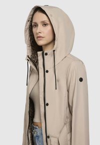 Beige waterdichte jas met een grote capuchon, zwarte hardware en een lederen voering, met een relaxte pasvorm en textuuraccenten.
