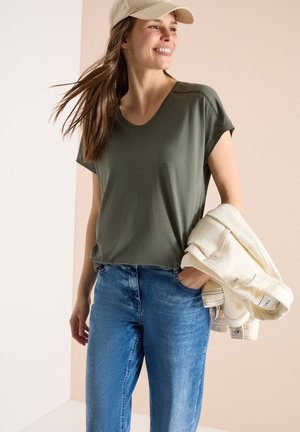 Femme souriante portant un t-shirt vert olive, une casquette beige, un jean bleu, tenant une veste légère pliée dans une main, posant devant un fond neutre.
