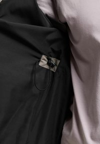 Schwarzer Stoff mit einem beigen Patch, der eine schwarze elastische Schnur und einen Verschluss hält, neben einer Person, die ein hellrosa Hemd trägt.