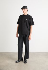 T-shirt nera con logo, taglio morbido, abbinata a pantaloni neri sartoriali e scarpe eleganti nere. Modello con un cappellino nero coordinato.