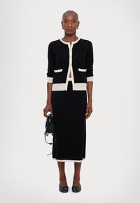 Black cardigan with white trim and pockets, paired with a matching black long skirt. Model holds a black fringed handbag.