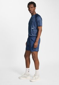 Marinblått idrottskläder i texturerad tyg; inkluderar t-shirt med kort ärm och shorts. Skorna är ljusgrå med vita detaljer och snören.