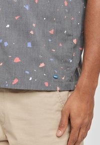 Maglietta a maniche corte grigia con un motivo maculato in rosso, rosa, blu e bianco. Presenta un'etichetta logo blu sull'orlo. Materiale in cotone o misto.