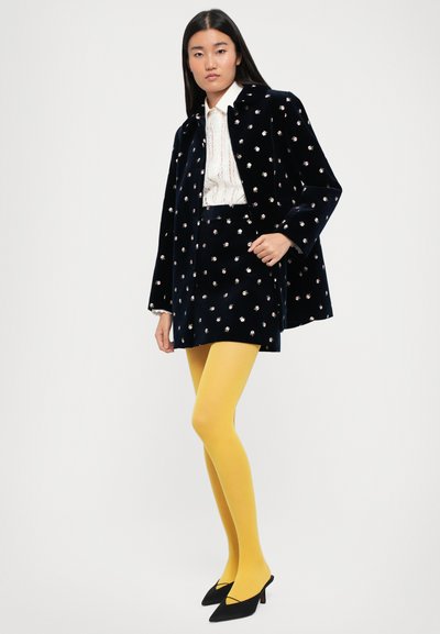 Tumesinise sametist mantel, millel on lillekirjad, koos valge tekstuuriga pluusi, tumesinise seeliku, erkoranžide sukkpükste ja mustade kontsakingadega.