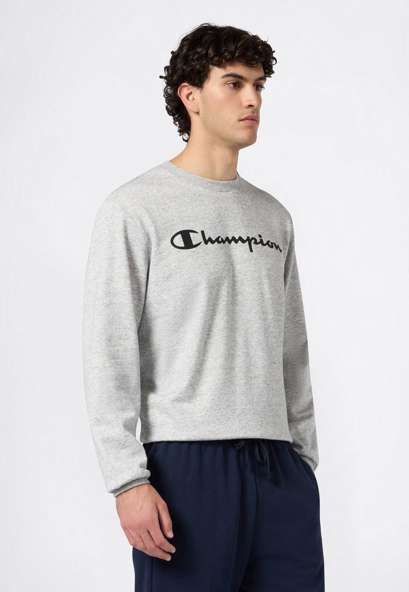 Sweat-shirt gris clair à manches longues avec logo "Champion" noir, col rond, poignets côtelés et coupe décontractée. Porté avec un pantalon à cordon de serrage bleu marine.