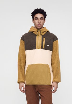 Jeune homme portant un sweat à capuche en polaire color-block avec des panneaux moutarde, marron et beige, debout devant un fond clair uni.