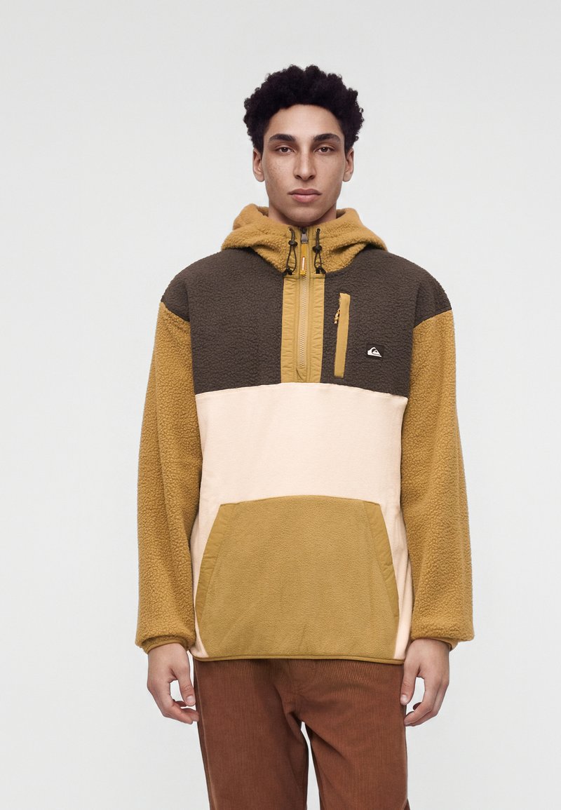 Νέος άνδρας με φούτερ fleece τύπου color-block σε μουσταρδί, καφέ και μπεζ αποχρώσεις, στέκεται μπροστά σε μια απλή ανοιχτή επιφάνεια.