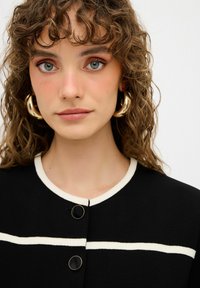 Prenda negra con rayas horizontales blancas, con un escote redondo y dos botones negros. El cabello rizado enmarca la cara.