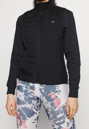 Person iført en sort lynlåsløs ærmeløs vest over en sort langærmet skjorte og multifarvede leggings med abstrakt mønster.