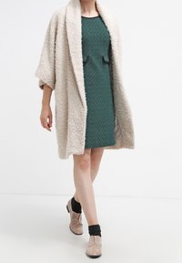 Beige, kuvioitu, avoin takki teal-värisen neulemekon päällä, jossa on nauhakiinnitys. Mallilla on päällään beige keng ät ja mustat nilkkasukat.