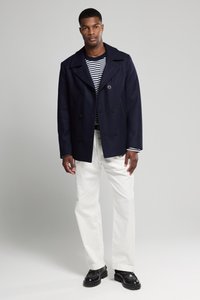 Granatowy wełniany płaszcz typu peacoat z podwójnym zapięciem, dużymi klapami i czarnymi guzikami, noszony na paskowanym podkoszulku, w połączeniu z białymi spodniami i czarnymi butami.