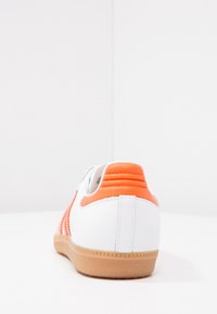 Vit sneaker med orange accenter, slätt lädermaterial, strukturerade detaljer på hälen och en sulan av gummi i naturfärg. Bilden visar baksidan.