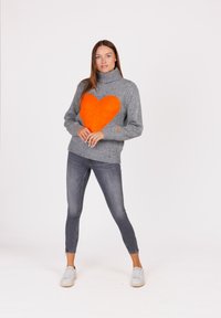 Vrouw staat met de handen gevouwen, gekleed in een grijze coltrui met een grote oranje hart en strakke grijze jeans met witte sneakers.