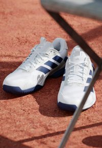 Chaussures de sport blanches avec des rayures bleues, dotées d'une tige en tissu texturé, d'une semelle en caoutchouc et d'un bout renforcé pour plus de durabilité.