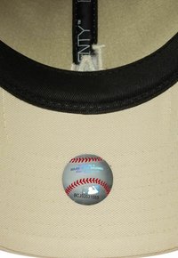 Helle beige Baseballkappe mit einem schwarzen Band innen. Auf der Unterseite befindet sich ein runder MLB-Aufkleber, der einem Baseball ähnelt.