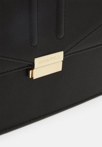 Borsa a tracolla nera in materiale sintetico con chiusura in metallo dorato e incisione "ANNA FIELD", superficie testurizzata e accenti cuciti verticalmente.