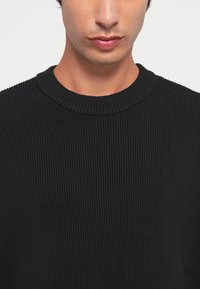 Svart ribbestrikket genser med rund hals, som har et teksturert mønster og en tettsittende design.