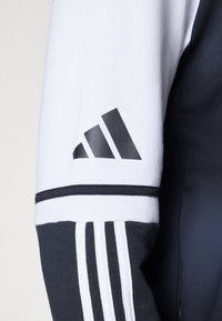 Veste de sport noir et blanc avec une manche supérieure texturée blanche et des accents noirs, trois rayures noires au niveau du poignet et un logo noir.