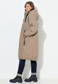 Lange khakifarbene äußere Jacke mit Kapuze, elastischem Saum und Reißverschlusstaschen. Getragen über dunklen Jeans und schwarzen Stiefeletten. Glatte Textur.