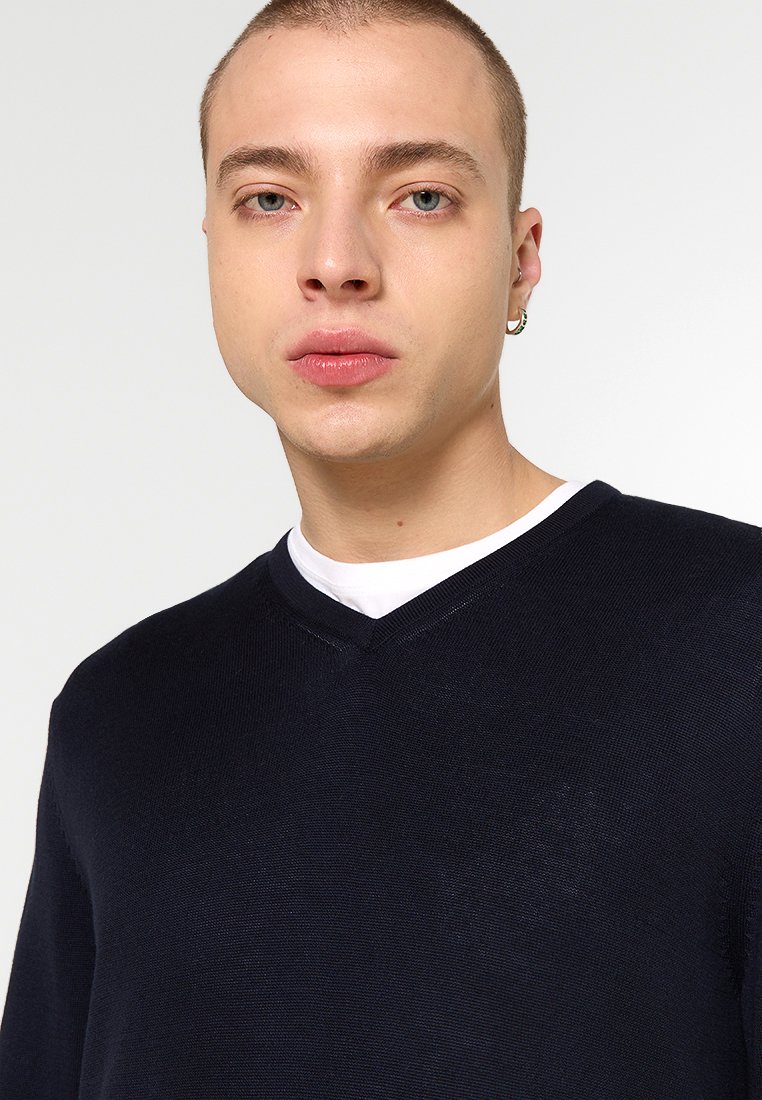 Marineblauer, gestrickter V-Ausschnitt-Pullover mit glatter Textur, subtilen horizontalen Mustern und über einem weißen T-Shirt getragen.