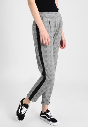 Personne portant un pantalon gris à carreaux avec une bande latérale noire et des baskets noires avec des lacets blancs, debout devant un fond uni.