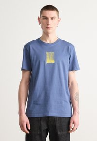 T-shirt de algodão azul com decote redondo e mangas curtas. Apresenta uma estampa abstrata amarela no centro e um design minimalista.