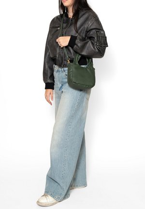 Vrouw draagt een zwarte leren jas en lichtblauwe wijd uitlopende jeans, met een groene handtas met schouderriem.