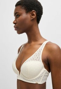 Light beige bra with a triangular cup shape, mesh fabric featuring a geometric pattern, and adjustable straps. Subtle brand accent at the side.
