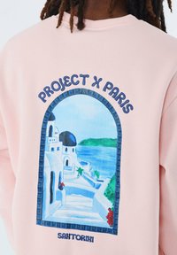 Sudadera rosa con un gráfico de Santorini, detalles arquitectónicos y acentos azules, con el texto "PROJECT X PARIS" arriba y "SANTORINI" abajo.