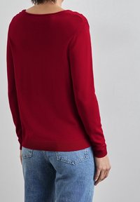 Person som bär en figurnära röd långärmad tröja och ljusblå denimjeans, visad bakifrån mot en enfärgad grå bakgrund.