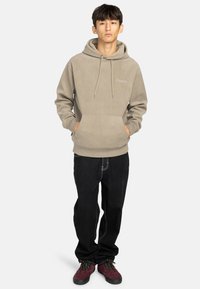 Sudadera beige con capucha, cordones y bolsillo frontal tipo canguro, combinada con jeans negros y zapatos burdeos. Textura de tela suave.