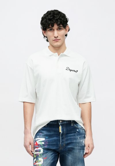 Camiseta polo blanca de manga corta con cuello y logo negro "Dsquared2". Combinada con jeans azules con patrones de salpicaduras de colores.