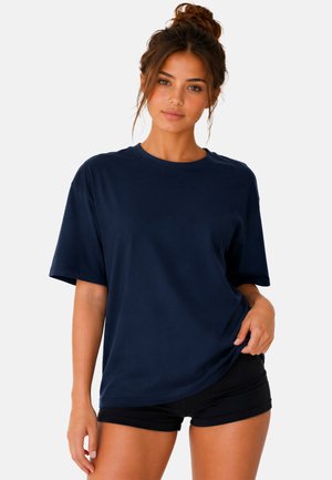 Junge Frau mit braunem, hochgebundenem Haar, die ein marineblaues übergroßes T-Shirt und schwarze Shorts trägt, steht vor einem schlichten hellen Hintergrund.