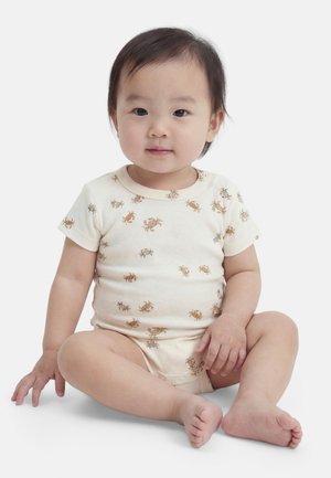 Dvojročné dieťa sedí s prekríženými nohami, oblečené v béžovom overalíku s malými vzormi krabov, hľadí dopredu s neutrálno výrazom.