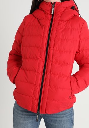 Rød quiltet pufferjakke med sort lynlås og hætte, båret med hænderne i lommerne, kombineret med blå jeans.