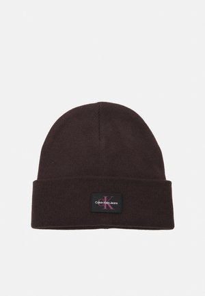 Bonnet en maille brun foncé avec revers comportant un patch noir rectangulaire marqué "Calvin Klein Jeans" en texte blanc et rouge.