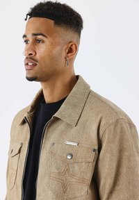 Chaqueta de tela color beige con cuello estructurado, cremallera frontal, dos bolsillos en el pecho y un sutil bordado de estrellas en hilo del mismo color.