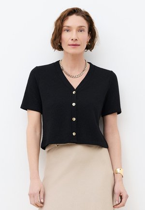 Vrouw met kort bruin haar, een zwart, kort mouwig topje met knopen, een beige rok, een gouden ketting, oorringen en een gouden armband.