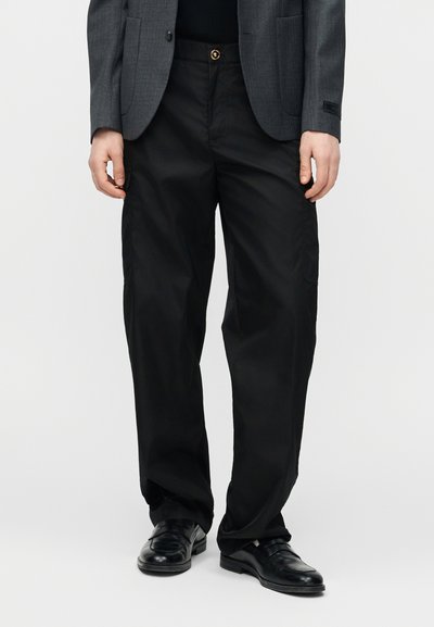 Personne portant un pantalon noir à jambes larges, des chaussures en cuir noires, un blazer gris foncé, debout devant un fond uni clair.