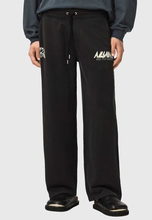 Person trägt lockere schwarze Jogginghosen mit weißem "ALLSAINTS"-Schriftzug und Grafiken, kombiniert mit schwarzen Schuhen und einem dunklen Langarmshirt.