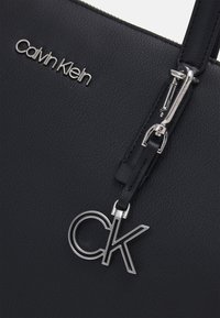 Sac à main en cuir noir arborant un logo "Calvin Klein" argenté et un charme amovible "CK" en argent, mettant en valeur une finition texturée et un design élégant.