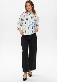 Camicia a maniche lunghe con colletto a fiocco, con una base bianca e un motivo geometrico in rosa, blu e verde. Abbinata a pantaloni neri a righe. 