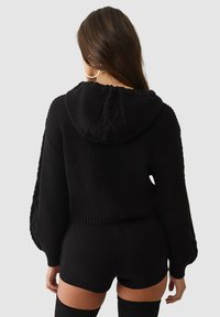 Schwarzer kurz geschnittener Hoodie mit gerippter Struktur, Kapuze mit Zopfmuster-Detail, kombiniert mit schwarzen Shorts. Modell von hinten gezeigt.