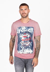 Roze crewneck T-shirt met een verouderde grafische print met tekst in het blauw en wit, tegen een donkerdere achtergrond. Korte mouwen, casual pasvorm.