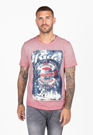Camiseta de cuello redondo rosa con un estampado gráfico desgastado que incluye texto en azul y blanco, sobre un fondo más oscuro. Manga corta, ajuste casual.