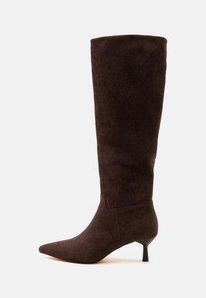 Botte en daim marron foncé montant au genou avec un bout pointu et un talon fin et bas, vue de profil sur un fond clair.