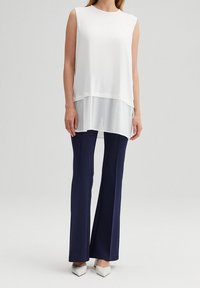 Witte mouwloze top met een geplooide onderkant, gecombineerd met marineblauwe broek met wijde pijpen en puntige witte schoenen. Glad materiaal en modern ontwerp.