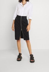 Svart denim blyertskjol med dragkedja fram, kombinerad med en vit skjorta med knappar och svarta sandaler med flera spännen och remmar.