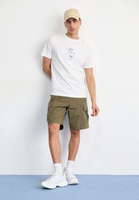 White short-sleeve T-shirt with a blue graphic, olive green cargo shorts, white sneakers, and a beige cap. Set against a plain background.